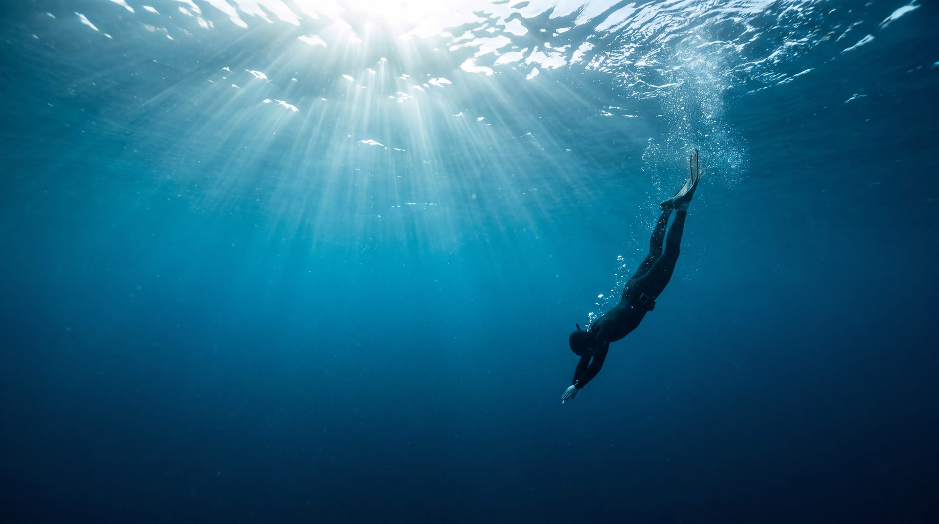 A freediver descending head-first through sunlit open water, trailing bubbles toward the surface
